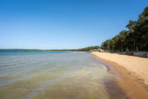 Varamobadets långsträckta sandstrand vid Vättern i Motala, med grunt klart vatten, gyllene sand och tallskog längs stranden under klarblå himmel.