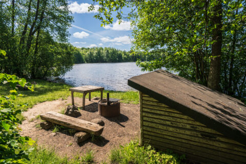 Vindskydd med grillplats och sittbänkar vid sjön i Västerby lövskogars naturreservat, omgivet av grönskande lövträd en solig sommardag