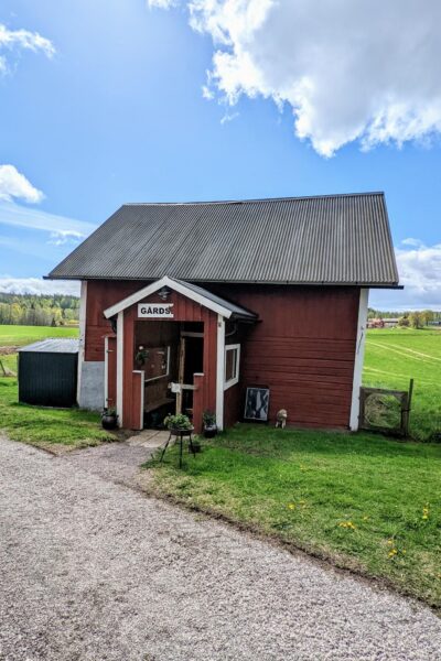 Röd gårdsbutik med skylt "Gårdsb" vid Brotorps Gård, omgiven av gröna åkrar och blå himmel i Östergötland.