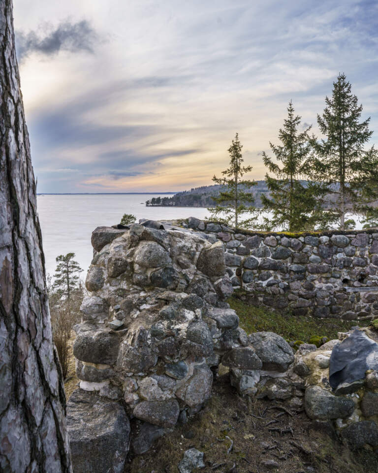 Ruinerna av Åls stenhus, en medeltida borg vid Roxens norra strand, med stenmurar omgivna av granar och utsikt över sjön i kvällsljus.