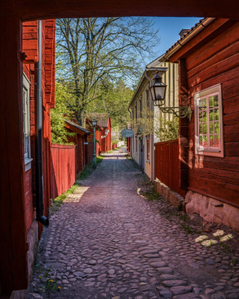 Kullerstenad gränd kantad av faluröda trähus och gula byggnader i Gamla Linköping, sedd genom en portöppning en solig vårdag