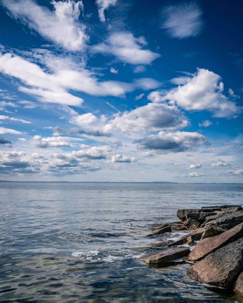 Klippor vid Borghamns strand med stilla vatten i Vättern under en blå himmel med vita moln