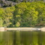 Stilla sjö med spegelblanka reflektioner vid badplatsen Norrkrog, med brygga, sandstrand och lummig lövskog mot bergig sluttning i Östergötland