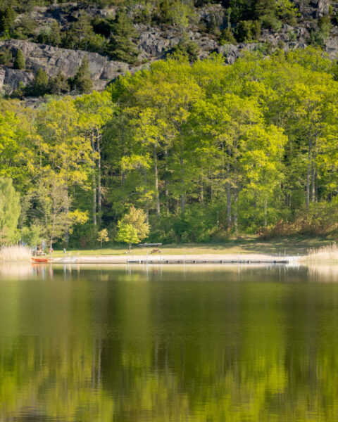 Stilla sjö med spegelblanka reflektioner vid badplatsen Norrkrog, med brygga, sandstrand och lummig lövskog mot bergig sluttning i Östergötland