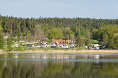 Campingplatser och röda stugor vid Stegeborgsgården i Norrkrog, speglade i det stilla vattnet i Slätbaken, omgivna av grön skog.