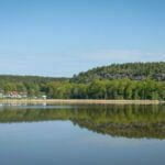 Spegelblank sjö vid Norrkrog med skogsklädda bergshöjder och Stegeborgsgårdens camping i bakgrunden, Östergötland