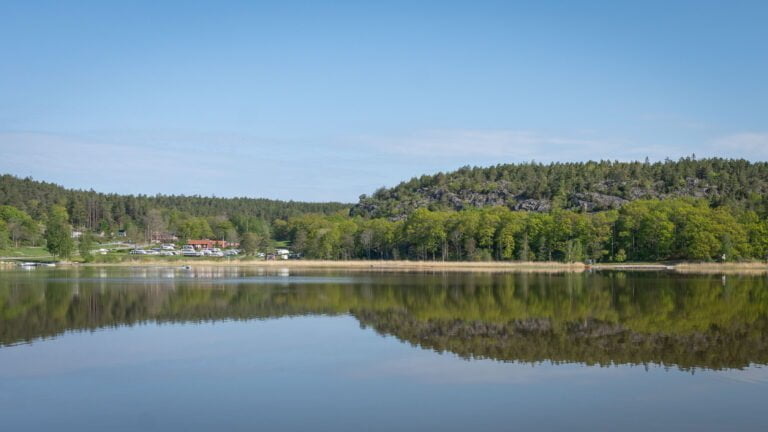 Spegelblank sjö vid Norrkrog med skogsklädda bergshöjder och Stegeborgsgårdens camping i bakgrunden, Östergötland