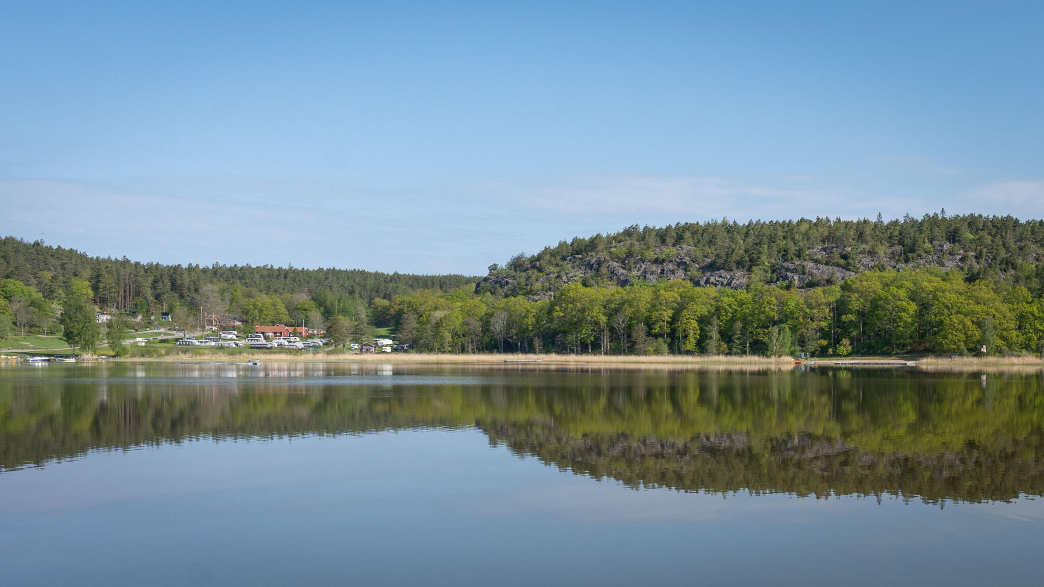 Spegelblank sjö vid Norrkrog med skogsklädda bergshöjder och Stegeborgsgårdens camping i bakgrunden, Östergötland