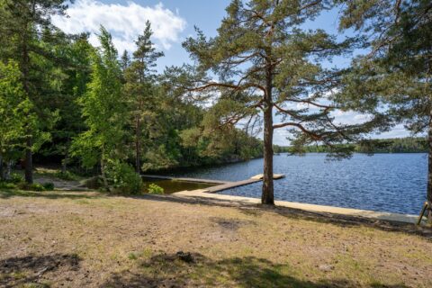 Sandstrand vid Gransjönäsbadet med en brygga som sträcker sig ut i sjön, omgiven av tallar och grön skog en solig sommardag.