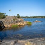 Klippor vid vattnet på Badholmarna i Östergötlands skärgård med en svensk flagga vajande mot klarblå himmel.