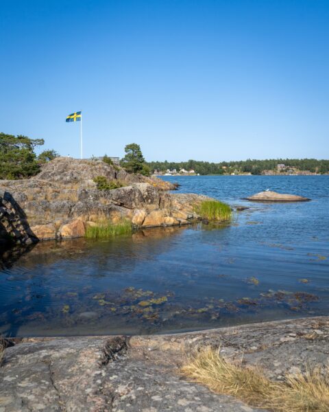Klippor vid vattnet på Badholmarna i Östergötlands skärgård med en svensk flagga vajande mot klarblå himmel.