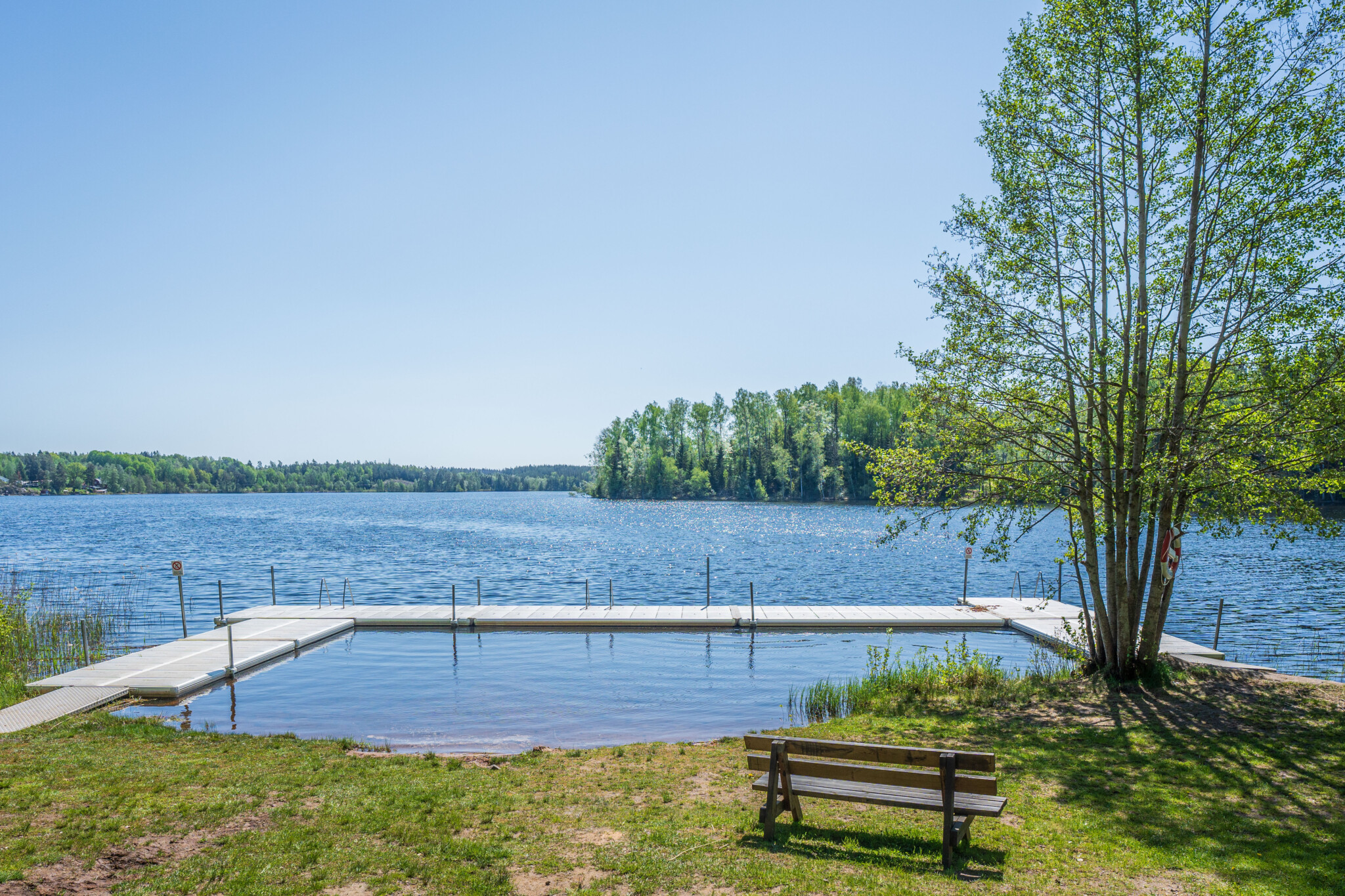 Bolens badplats vid Simonstorp med avskärmad barnbassäng, brygga, parkbänk och utsikt över sjön en solig sommardag.