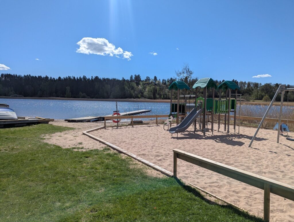 Lekplats med rutschkana och gungor på sandstrand vid Dalbystrand, med badbrygga och skogsklädd strand i bakgrunden.