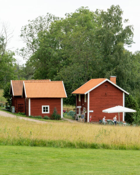 Röda faluröda timmerstugor med tegeltak vid Farfarstugan café i Övralid, Motala, med uteservering under parasoll och gröna ängar framför.
