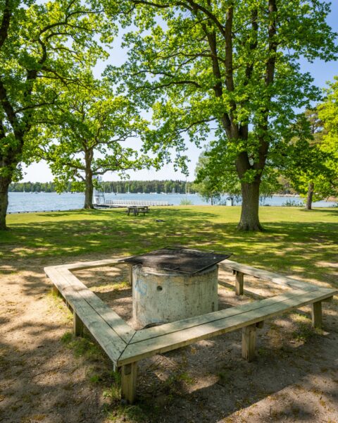 Grillplats med sittbänkar under lövträd vid Råssnäsbadet i Motala, med grön gräsmatta och sjöutsikt mot Vättern.