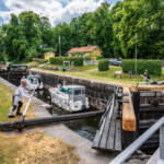 Fritidsbåtar slussas genom Nykvarns sluss vid Göta kanal, en man manövrerar slussportens arm medan gula hus och grönska omger kanalen.