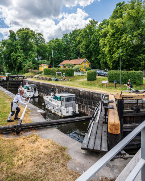 Fritidsbåtar slussas genom Nykvarns sluss vid Göta kanal, en man manövrerar slussportens arm medan gula hus och grönska omger kanalen.