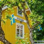 Gult trähus med skylten "Nykvarn" och svenska flaggor, omgivet av grönskande träd vid Nykvarns sluss i Linköping.