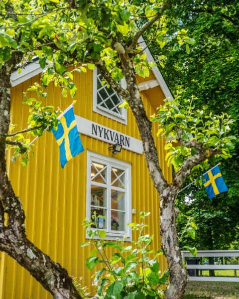 Gult trähus med skylten "Nykvarn" och svenska flaggor, omgivet av grönskande träd vid Nykvarns sluss i Linköping.