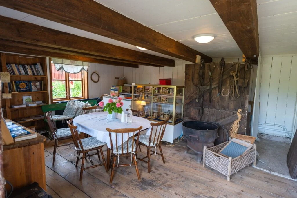 Rustikt rum i Sjölingården med träbjälkar, brädgolv, matbord med blommor, gamla redskap på väggen och en utställningsmonter med föremål.