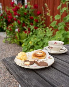 Svensk fika med kanelbulle, kakor och kaffekopp på träbord utanför Sjölingården, med röda rosor och faluröd vägg i bakgrunden.
