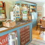 Gammaldags specerihandel i Sjölingården med blåmålad disk, vykortställ, mjölkkruka och skylt med texten "Speceri-Handel" på trägolv med trasmattor.