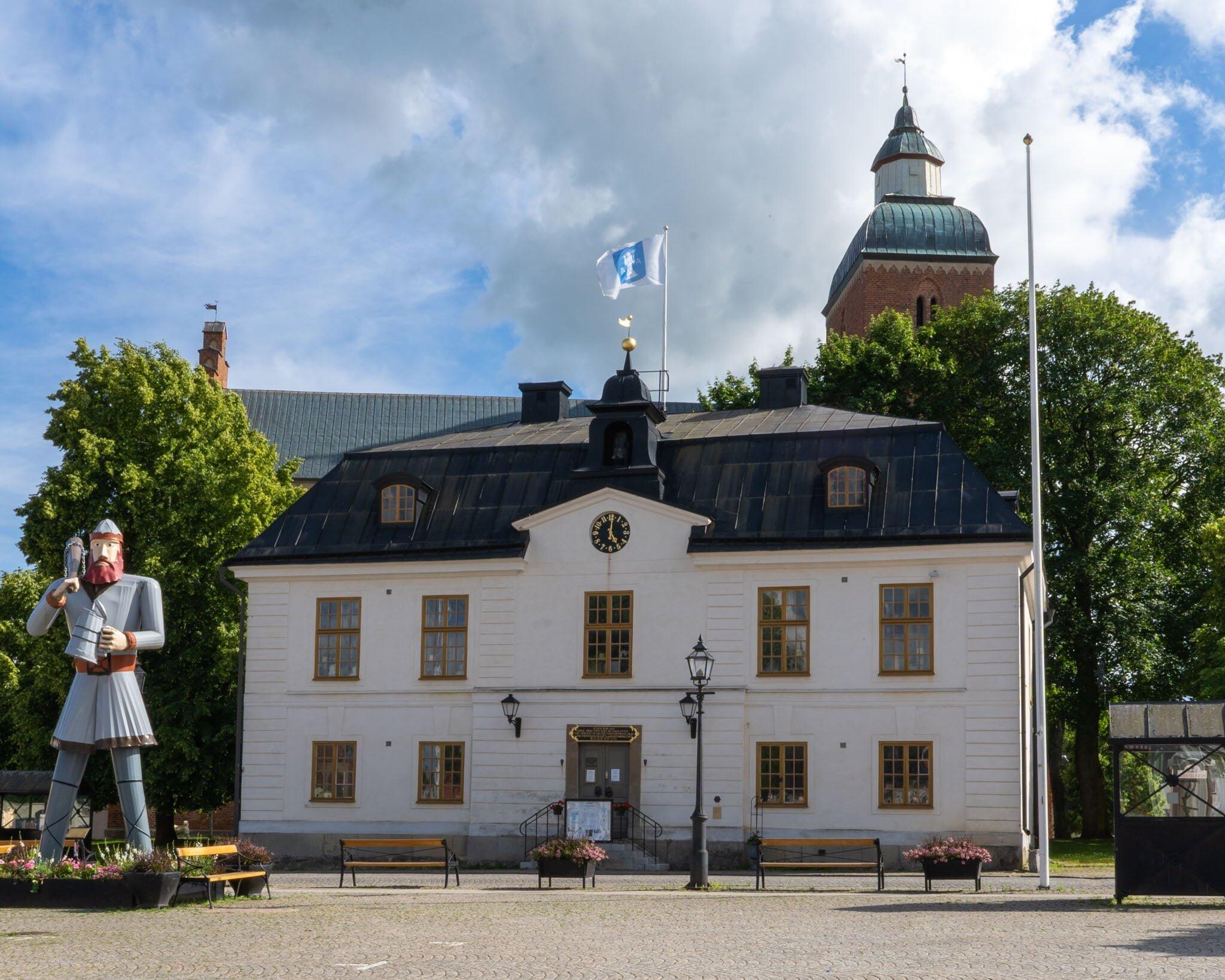 Skänninge rådhus med vit fasad och klocka, omgivet av gröna träd, med en riddarskulptur till vänster och kyrkans kupol i bakgrunden.