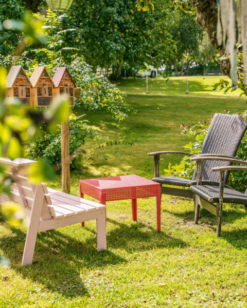 Mysig trädgårdshörna med rosa bänk, rött bord, flätad stol och insektshotell omgivet av grönskande gräsmatta och fruktträd i Borensberg, Östergötland.