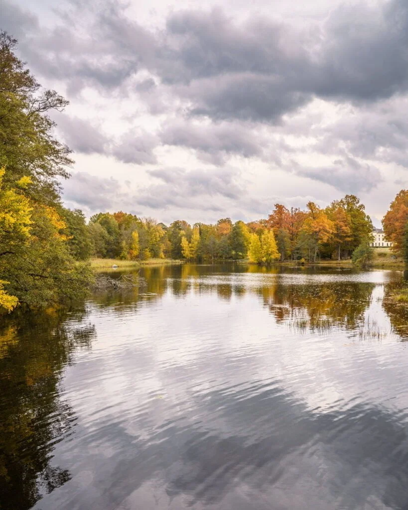 Stilla sjö omgiven av höstfärgade lövträd i gult, orange och grönt vid Bjärka-Säby, med dramatisk molnhimmel och speglingar i vattnet