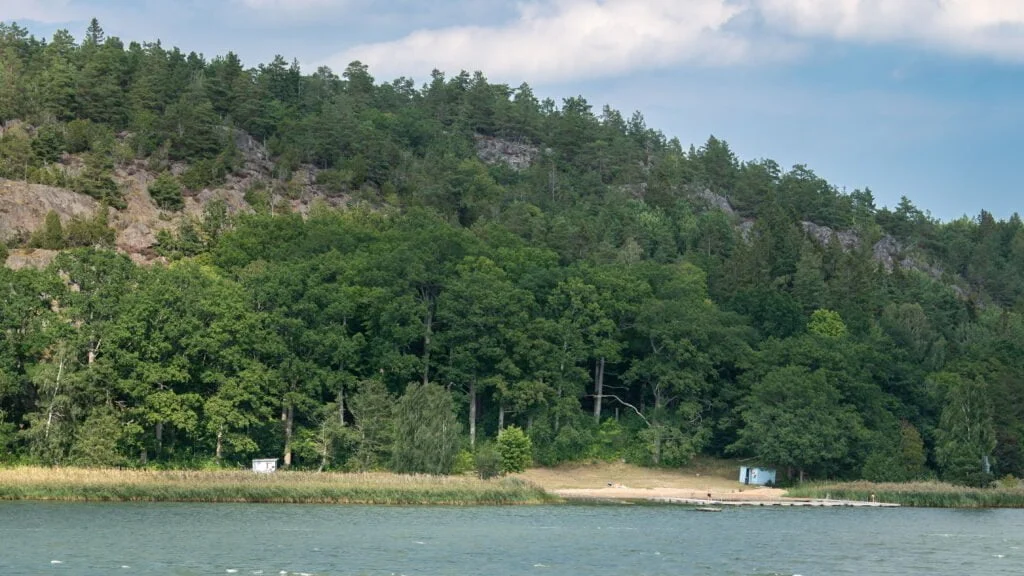 Sandstrand vid Stegeborgsgårdens badplats med lugnt vatten och en skogsklädd bergsförkastning i Norrkrogs naturreservat, Östergötland