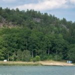 Sandstrand vid Stegeborgsgårdens badplats med lugnt vatten och en skogsklädd bergsförkastning i Norrkrogs naturreservat, Östergötland