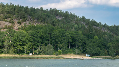 Sandstrand vid Stegeborgsgårdens badplats med lugnt vatten och en skogsklädd bergsförkastning i Norrkrogs naturreservat, Östergötland