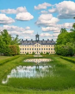 Sturefors slott med gul fasad och klocktorn, speglat i en damm omgiven av gröna gräsmattor och lummiga träd en sommardag.