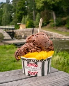 Glassbägare från Sänkdalens Glass med choklad- och mangoglass på ett träbord vid Brådtom sluss längs Göta kanal, omgivet av grönska.