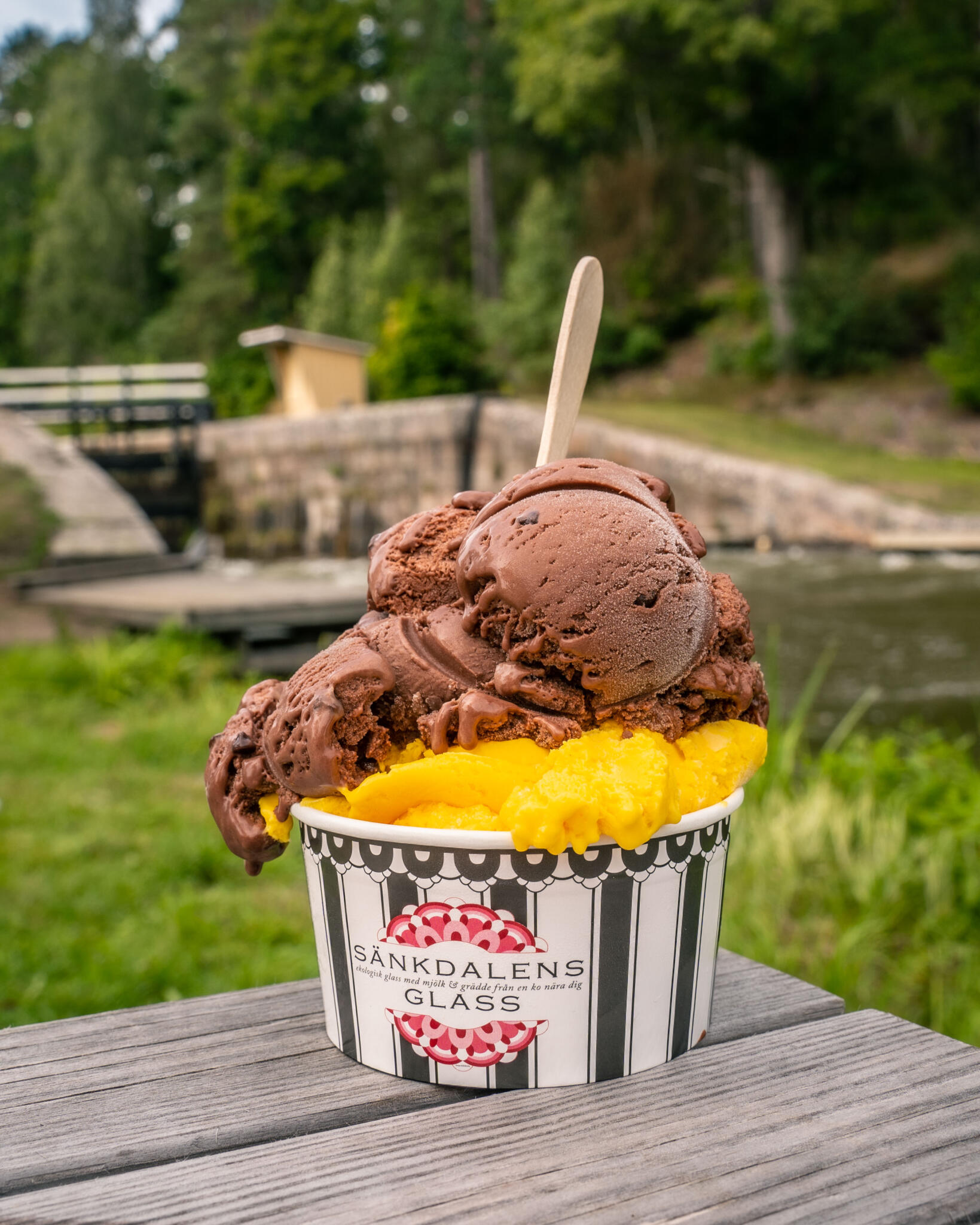 Glassbägare från Sänkdalens Glass med choklad- och mangoglass på ett träbord vid Brådtom sluss längs Göta kanal, omgivet av grönska.