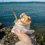 Hand som håller en bägare glass från Kungens glass vid Vätterns strandkant i Vadstena, med blått vatten och klart himmel i bakgrunden