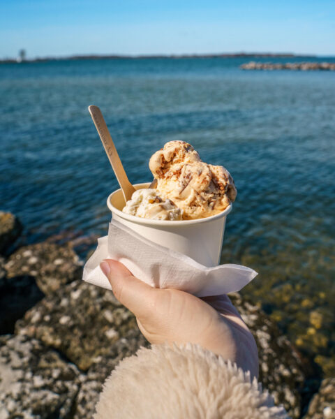 Hand som håller en bägare glass från Kungens glass vid Vätterns strandkant i Vadstena, med blått vatten och klart himmel i bakgrunden