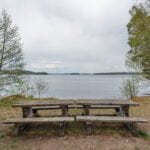 Två rustika träbänkar med bord vid vattnet i Hallstad Ängar med utsikt över sjön, Kinda kommun i Östergötland