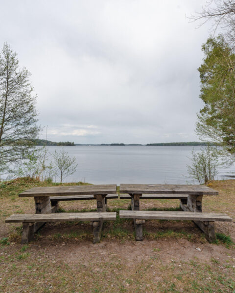 Två rustika träbänkar med bord vid vattnet i Hallstad Ängar med utsikt över sjön, Kinda kommun i Östergötland