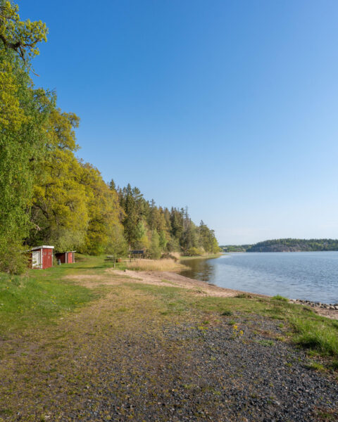Husbyvik badplats vid Slätbaken nära Mogata med sandstrand, litet rött omklädningshus, grönskande lövträd och stilla vatten en solig vårdag.