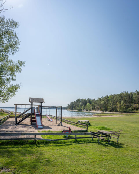 Lekplats med rutschkana och gungor vid Mon badplats i Sankt Anna skärgård, med gröna gräsytor, havsvik och tallskog i bakgrunden.