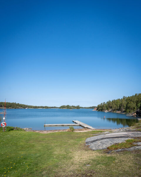 Badbrygga vid Tyrislöts badplats i Sankt Annas skärgård med stilla vatten, grönska, klippor och livboj en solig sommardag.