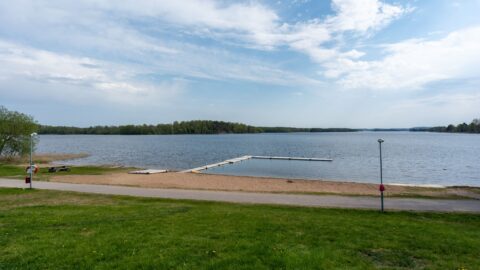 Åsundabadet vid Åsunden i Kinda kommun med sandstrand, badbrygga i L-form, gröna gräsytor och lugnt vatten en molnig vårdag.
