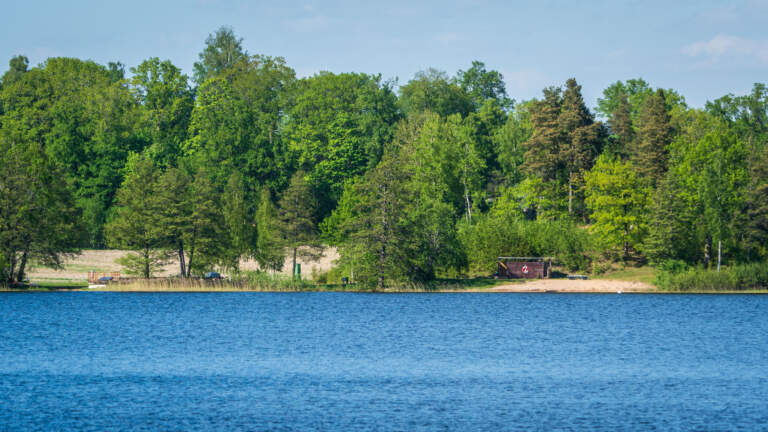 Byle badplats vid sjön Tisnaren med sandstrand, omklädningshytt och grönskande lövträd en solig sommardag i Finspångs kommun