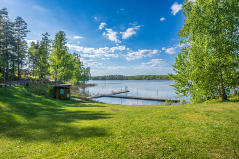 Grön gräsmatta vid Hävla badplats med badbrygga i sjön Tisnaren, omgiven av lövträd och tallar en solig sommardag.