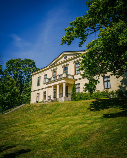 Charlottenborgs slott i Motala kommun, en gulvit klassicistisk herrgårdsbyggnad med pelare och balkong omgiven av gröna träd och gräsmatta en solig dag.
