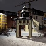 Snötäckt lekplats i Mjölnareparken i Norrköping med miniatyrbyggnader, bland annat ett klocktorn märkt 1750 och ett gult hus, upplysta en vinterkväll.