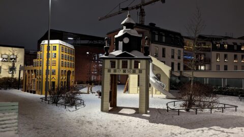 Snötäckt lekplats i Mjölnareparken i Norrköping med miniatyrbyggnader, bland annat ett klocktorn märkt 1750 och ett gult hus, upplysta en vinterkväll.