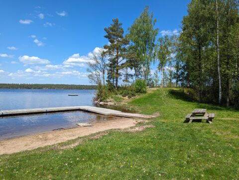 Sandstrand och brygga vid Rejmyre badplats vid sjön Hunn, med picknicbord på gröna gräsytor, björkar och blå himmel en sommardag