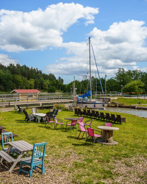 Uteservering vid Jollas Trädgårdskafé vid Göta kanal med färgglada stolar, bord på gräsmatta och en segelbåt i slussen.